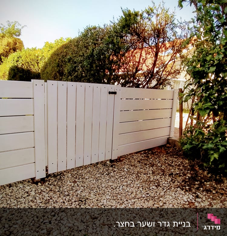 גדר עץ לבנה על חצץ ליד צמחייה ירוקה